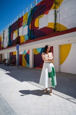 Şık bir kadın canlı, geometrik boyanmış bir duvara karşı güvenle poz veriyor, şehir modasını ve renkli sokak sanatını sergiliyor.