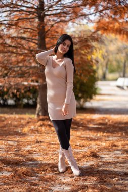 Sonbahar boyunca parkta poz veren genç bir kadın, bej bir elbise ve yerde yaprakları olan kovboy çizmeleri giyiyor.