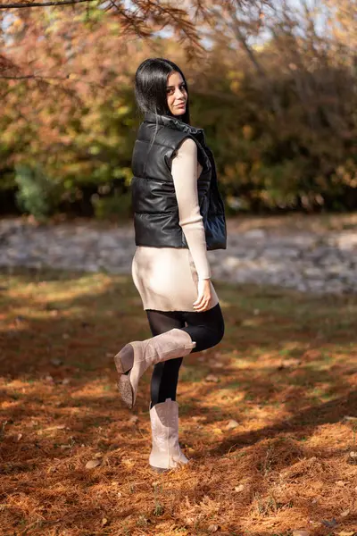 Kovboy çizmeleri giyen, siyah yelek ve bej elbiseli genç bir kadın parkta sonbahar renklerinin tadını çıkarıyor.