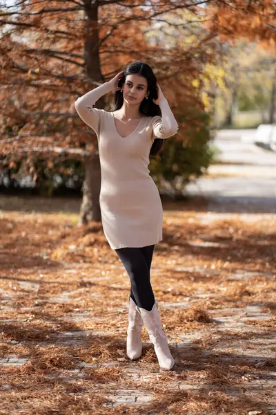 Sonbahar parkında bej elbise ve kovboy çizmeleri giyerek poz veren manken.