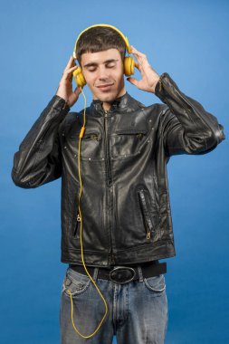 Deri ceket giyen genç adam sarı kulaklıklı müzik dinliyor, kapalı gözlerle sesin tadını çıkarıyor.