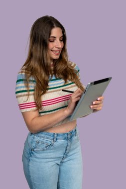 Stylus kalemle tablet kullanan gülümseyen genç kadın, mor bir arka plan üzerinde çalışıyor ya da çalışıyor.
