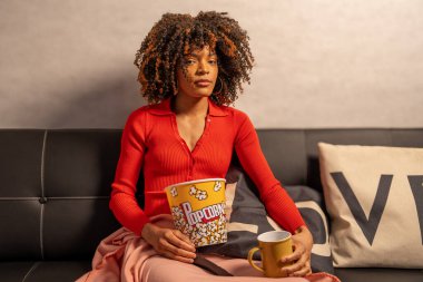 Kanepede oturan genç kadın elinde patlamış mısır kovası ve kahve bardağıyla evde film gecesinin tadını çıkarıyor.