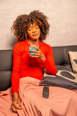 Hamile bir kadın koltukta rahat bir akşamın tadını çıkarıyor, canlı bir mavi kokteyl içiyor ve televizyon seyrediyor.