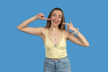 Neşeli genç bir kadın canlı mavi bir arka planda gülümserken yanaklarını gösteriyor.