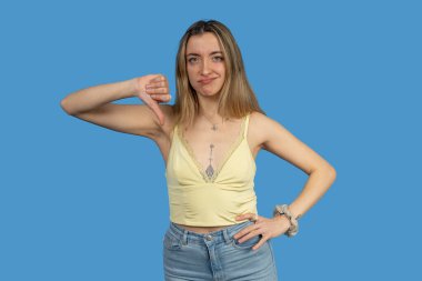 Onaylamadığını belirten genç bir kadın. Baş parmağıyla işaret ediyor. Stüdyo resmi mavi arka planda.