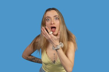Genç sarışın kadın şaşırmış görünüyor, elleri ağızda, jest yapıyor ve mavi arka planda şaşkınlık var.
