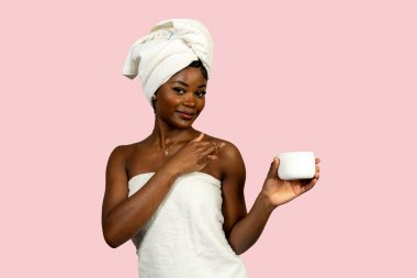 Havluya sarılı gülümseyen genç kadın duş aldıktan sonra vücut kremi sürüyor.