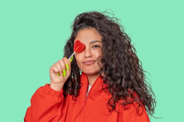 Gözleri üzerinde kalp şeklinde bir lolipop tutan kıvırcık saçlı, tatlı ve yaramaz bir tavır sergileyen neşeli genç bir kadın.