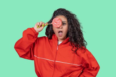 Stüdyoda genç bir kadının gözünün üstüne lolipop tutarken ve yeşil bir arka plana bakarken görüntüsü.