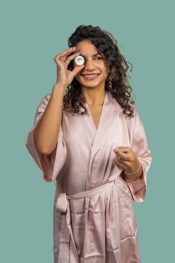 Bornoz giymiş genç bir kadın elinde nemlendirici krem kavanozuyla güzellik ürünlerini tanıtıyor.