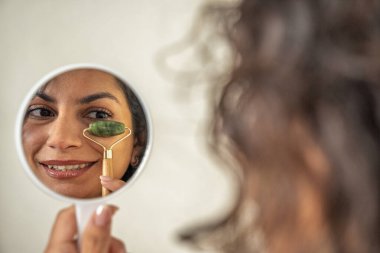 Yüzüne masaj yapan yeşim bir silindir kullanan gülümseyen kadın küçük bir aynaya yansıyor.