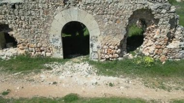 Parçalanmış taş duvarlar ve terk edilmiş bir kalenin kemerli girişi, üzerinde grafiti izleri olan, aşırı gelişmiş bitki örtüsünün ortasında duruyor.