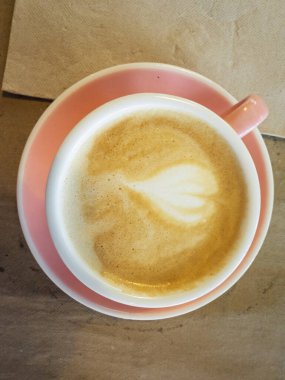 Kahve fincanı ve latte sanat kalp tasarımı pembe çay tabağı ve kahverengi kağıt tabak altlığı üzerine oturur.