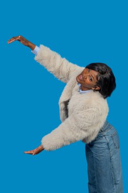 Zarif genç bir kadın, elleri açık hareket ediyor, parlak mavi bir arka planda bir şeyler sunuyor.