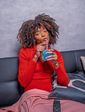 Kıvırcık saçlı genç bir kadın rahat bir kanepede dinlenirken ferahlatıcı mavi bir kokteylin tadını çıkarıyor.