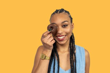 Stüdyo portresi, uzun örgülü, çikolatalı çörekleri gözünün üstünde tutan, canlı sarı arkaplana karşı tatlı ikramlardan hoşlanan, gülümseyen genç bir kadın.