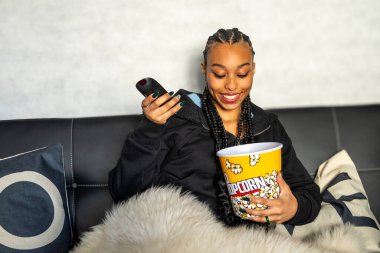 Uzaktan kumandalı ve patlamış mısır kovalı gülümseyen kadın, evde rahatlatıcı bir film gecesinin tadını çıkarıyor.