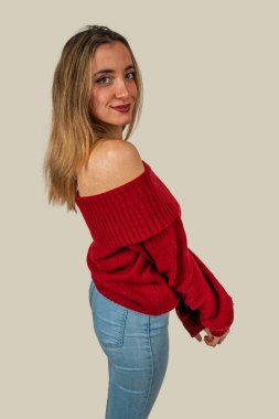 Uzun sarı saçlı, omuzları açık kırmızı bir kazak ve kot giyen genç bir kadının stüdyo portresi.