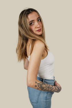 Uzun sarı saçlı ve dövmeli genç bir kadın stüdyoda poz veriyor, beyaz bluz ve kot pantolon giyiyor.