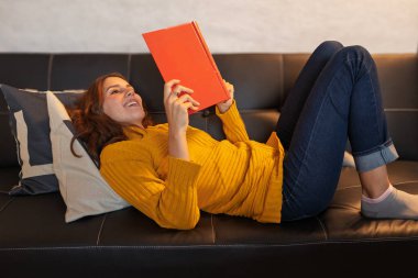 Kanepede uzanmış turuncu bir kitap okumaktan zevk alan genç bir kadın edebiyatta rahatlık ve eğlence buluyor.