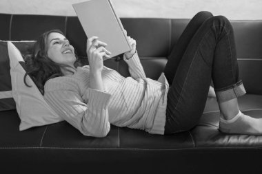 Genç bir kadın kanepede uzanıp kitap okuyor, evde rahatlatıcı bir anın tadını çıkarıyor.