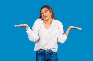 Kafa karışıklığı ve belirsizliği ifade eden genç bir kadın, açık avuçlarla omuzlarını silkeliyor canlı mavi bir arka plana karşı.