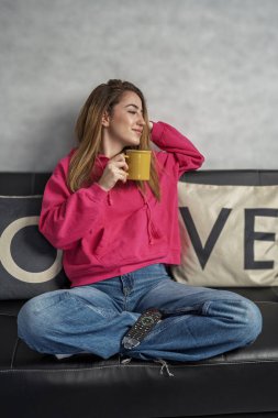 Huzurlu genç kadın rahat koltuğunda dinleniyor, sıcak bir içeceğin tadını çıkarıyor ve elinde uzaktan kumandayla televizyon seyrediyor.