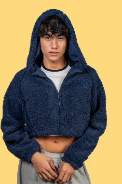 Modaya uygun genç adam mavi kürklü kapüşonlu ve gri eşofmanlı, sarı arka planda poz veriyor.
