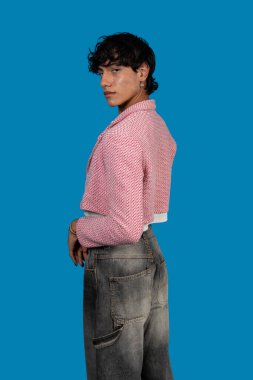 Pembe kesilmiş ceket ve mavi arka planda gri kot pantolon giyerken omuzlarının üzerinden bakan modaya uygun genç latin model.