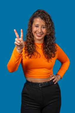 Mavi arka planda zafer işareti gösteren gülümseyen genç bir kadının stüdyo portresi