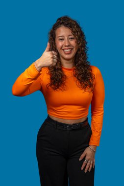 Neşeli genç bir kadının stüdyo portresi. Parmakla işaret ediyor, onay veriyor ve canlı mavi arkaplana karşı olumlu tavır sergiliyor.