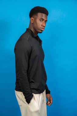 Uzun kollu siyah gömlek ve mavi arka planda bej pantolon giyen genç bir adamın stüdyo fotoğrafı.