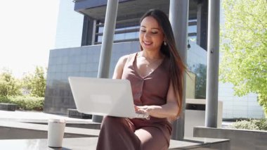 Modern ofis binasının dışında dizüstü bilgisayar kullanan genç bir iş kadını.