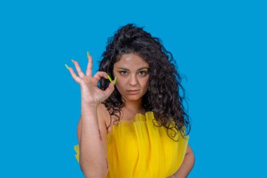 Kendine güveni tam genç bir kadın yeşil akrilik tırnaklı iyi bir jest yapıyor. Onayını ve anlaşmasını canlı mavi arka planda dile getiriyor.