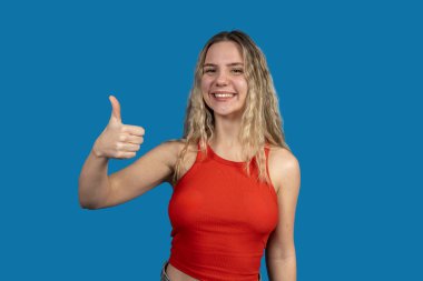 Gülümseyen genç bir kadın başparmak işareti yapıyor, pozitif ifade ediyor ve mavi bir zemin üzerinde mutabakat gösteriyor.