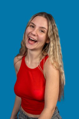 Uzun sarı saçlı, kırmızı bluzlu ve kotlu neşeli genç bir kadının portresi, canlı mavi bir arka plana yürekten gülüyor.