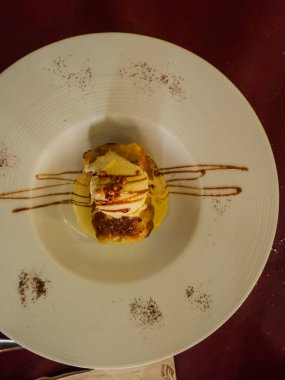 Beyaz bir tabakta nefis bir tatlı, içinde dondurma, karamel yağmuru, çikolata parçacıkları ve muhallebi var, görsel olarak çekici bir mutfak deneyimi yaratıyor.