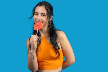 Kırmızı bir lolipopun tadını çıkaran genç bir kadının canlı bir mavi arkaplana karşı çekimleri.