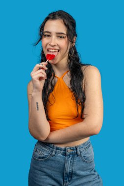 Kırmızı kalp şeklindeki lolipopun tadını çıkaran genç bir kadının canlı mavi bir arka planda çekimleri.