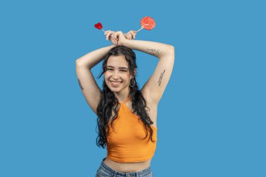 Elinde iki lolipop tutan neşeli genç bir kadın, biri kalp şeklinde, diğeri yuvarlak, parlak mavi bir arka planda.