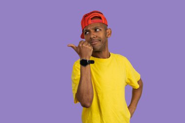 Stüdyo portresi, sarı tişört ve kırmızı beysbol şapkası giyen, başparmağıyla ve yana bakan, mor arka planda şüphe ve şüpheyi ifade eden genç bir adam.
