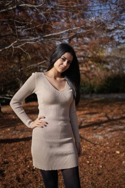 Dışarıda poz veren genç bir kadın, bir sonbahar parkında çıplak ağaçlar ve düşmüş yapraklarla duruyor.