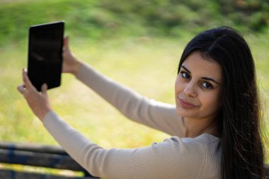 Gülümseyen, elinde dijital tabletle dışarıda çekilmiş bir selfie videosu ya da resmi çeken bir kadın.
