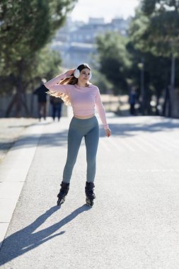 Güneşli bir günde parkta kulaklıkla paten kayan genç bir kadın.