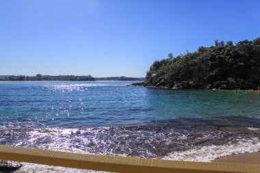 Berrak mavi gökyüzü, parlak okyanus dalgaları ve kayalık kıyılarda yemyeşil bitki örtüsüyle sakin bir sahil manzarası. Kıyı parlak güneş ışığıyla aydınlanır..