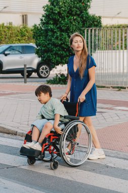 Anne, oğlunun tekerlekli sandalyesini iterek karşıya geçiyor. Yaşam tarzı konsepti.