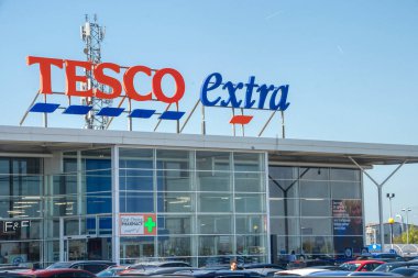 Binanın tepesinde koyu kırmızı ve mavi harflerle yazılmış Tesco Extra tabelası olan cam önlü süpermarket girişi..