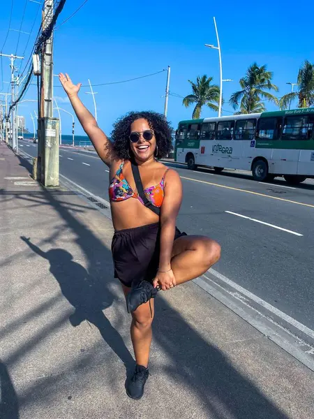 Renkli bir bikini giyen kıvırcık saçlı ve güneş gözlüklü genç bir kadın, arka planda palmiye ağaçları ve halk otobüsleriyle deniz kenarında yürürken neşeli bir poz veriyor. O gülümserken gün ışığı parlıyor, rahatlamasını yansıtıyor.