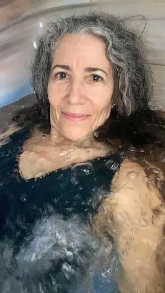 Gri saçlı, gülümseyen bir kadın karanlık suyla dolu bir küvette dinleniyor. Yumuşak ışık, onun sakin ifadesini ve etrafındaki kabarcıkları vurguluyor. Huzur ve refahı çağrıştırıyor..